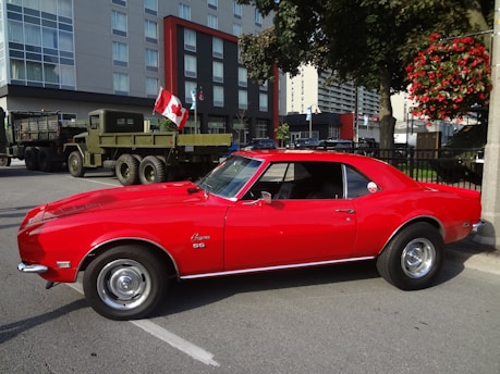 A bright red vintage sports car is parked on a city street. The car's sleek design features prominent curves and shining rims. Behind the car, a military truck is parked, displaying a Canadian flag. The backdrop includes a modern multi-story building, indicating an urban setting. Greenery, including a tree and a hanging flower basket, adds color to the scene.