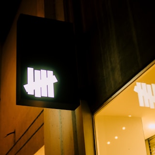 An illuminated sign with a white tally mark design consisting of five vertical lines. The sign is attached to the exterior of a building, which appears to have a modern design with clean lines and a mixture of dark and light surfaces. The sign's reflection is visible in a nearby window.