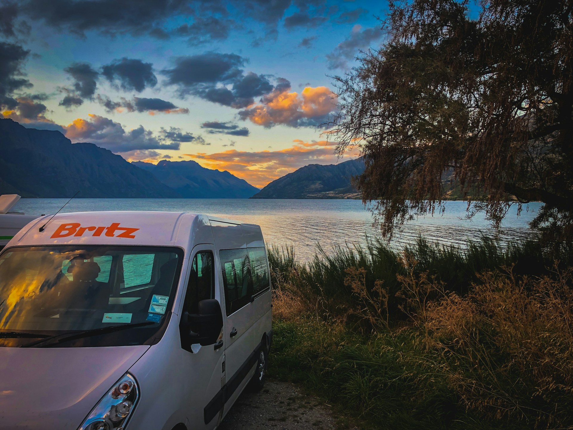 A rugged off-road caravan parked beside a serene Australian outback lake at sunset, with Mr Jim and Wrong Way Wendy setting up camp.