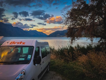 A serene lakeside scene with a camper van parked near the edge of a body of water, surrounded by tall grass and trees. The sky exhibits a vibrant sunset with hues of orange and blue, with mountains in the distance.