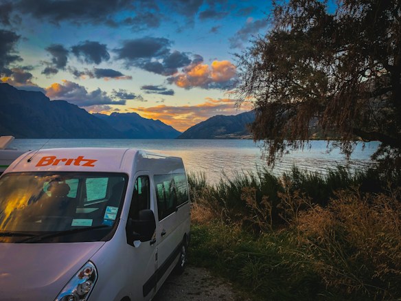 A serene lakeside scene with a camper van parked near the edge of a body of water, surrounded by tall grass and trees. The sky exhibits a vibrant sunset with hues of orange and blue, with mountains in the distance.