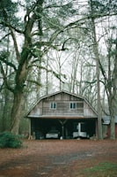 A rustic wooden barn with a slightly weathered appearance is nestled among tall trees. The surrounding trees are bare, suggesting a fall or early winter setting. The barn has a metal roof and is partly open at the front, revealing an old vehicle and some equipment stored inside. The ground is covered with fallen leaves and the color palette is muted with earthy tones.