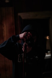 A person wearing a dark hoodie is holding a camera up to their face, capturing an image. The setting appears to be dimly lit, with wooden panels visible in the background. The person is partially obscured by shadows, creating a moody atmosphere.