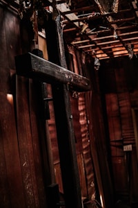 A large wooden cross is leaning against an old wooden wall, surrounded by a dimly lit and rustic room with exposed beams and wooden panels. Organic materials hang from the ceiling, and the space appears aged and weathered.