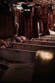 An abandoned interior space with wooden pews in disrepair, covered in dust and debris. The walls and ceiling show signs of neglect with exposed wooden beams and broken panels. Dim lighting adds to the somber atmosphere.