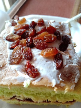 A slice of layered cake with a light green interior, topped with white frosting and sprinkled with golden and dark brown raisins. The cake appears moist and fluffy with visible textures in the frosting.