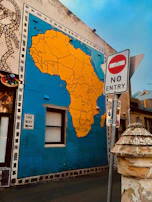 A large mural of Africa painted on a building wall with bright yellow and blue colors. Nearby, there is a 'No Entry' road sign and a 'One Way' sign attached to the wall. The surrounding area has urban elements such as a street lamp and textured walls.