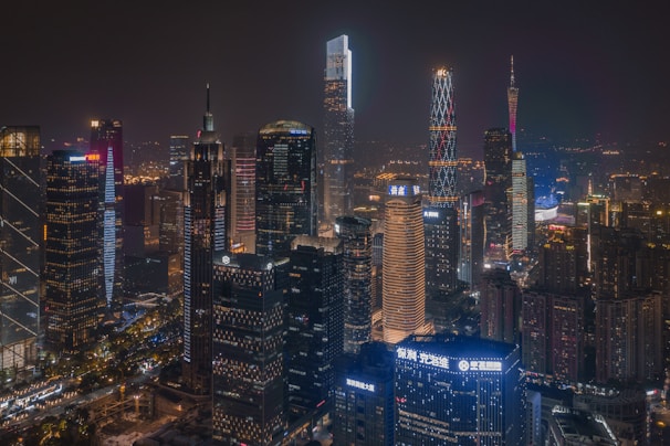 A nighttime cityscape of a sprawling metropolis with a dense cluster of modern skyscrapers illuminated by vibrant lights. The skyline is dominated by tall buildings with diverse architectural styles, featuring brightly lit signs and colorful lighting displays. The scene captures the bustling urban environment with vehicles and streets visible below.