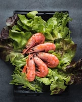 A black rectangular plate holds a fresh arrangement of shrimp on a bed of mixed green lettuce leaves. The shrimp are pink and appear to be cooked, garnished with small herb flecks. The vibrant greens of the lettuce contrast with the shrimp's color.
