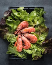 A black rectangular plate holds a fresh arrangement of shrimp on a bed of mixed green lettuce leaves. The shrimp are pink and appear to be cooked, garnished with small herb flecks. The vibrant greens of the lettuce contrast with the shrimp's color.
