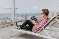 A person wearing sunglasses is lounging on a white deck chair on a balcony, overlooking a coastal view. They are casually dressed in a red plaid shirt and black jeans, and they are holding and reading a book titled 'Expert Secrets'. The ocean can be seen in the background along with palm trees and buildings.