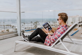 A person wearing sunglasses is lounging on a white deck chair on a balcony, overlooking a coastal view. They are casually dressed in a red plaid shirt and black jeans, and they are holding and reading a book titled 'Expert Secrets'. The ocean can be seen in the background along with palm trees and buildings.