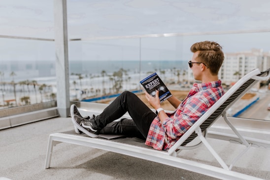 A person wearing sunglasses is lounging on a white deck chair on a balcony, overlooking a coastal view. They are casually dressed in a red plaid shirt and black jeans, and they are holding and reading a book titled 'Expert Secrets'. The ocean can be seen in the background along with palm trees and buildings.