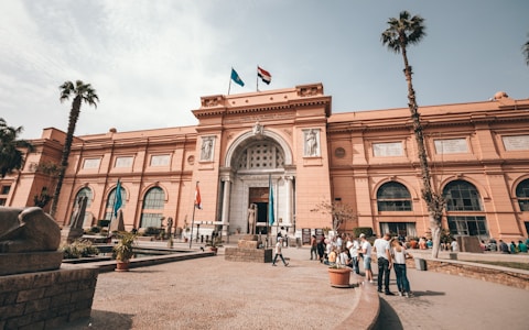 A large, historic building with a reddish-brown facade, adorned with statues and arched windows. Palm trees line the front, and people are gathered near the entrance. Two flags fly atop the building.