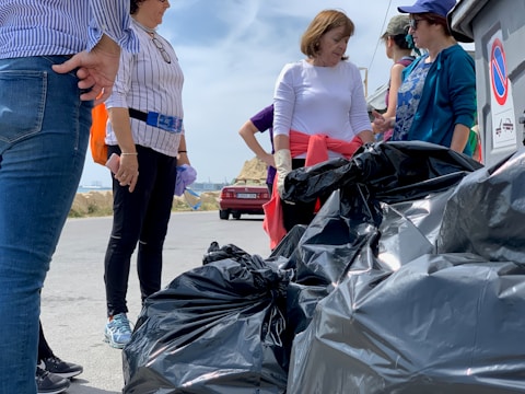 A group of people is gathered outdoors, participating in what appears to be a clean-up activity. They are surrounded by large black garbage bags. The people are wearing casual clothing and some have gloves on their hands.