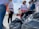 A group of people is gathered outdoors, participating in what appears to be a clean-up activity. They are surrounded by large black garbage bags. The people are wearing casual clothing and some have gloves on their hands.