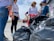 A group of people is gathered outdoors, participating in what appears to be a clean-up activity. They are surrounded by large black garbage bags. The people are wearing casual clothing and some have gloves on their hands.
