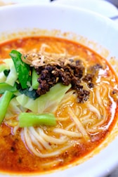 A bowl of spicy noodle soup topped with minced meat, fresh green vegetables, and crispy fried onions. The soup base is rich and reddish-orange, indicating a flavorful and possibly spicy broth.