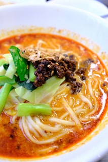 A bowl of spicy noodle soup topped with minced meat, fresh green vegetables, and crispy fried onions. The soup base is rich and reddish-orange, indicating a flavorful and possibly spicy broth.