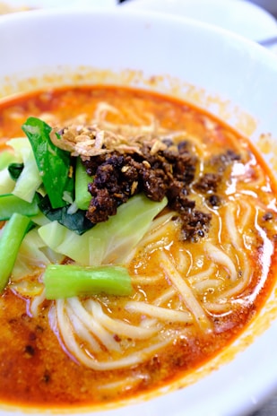 A bowl of spicy noodle soup topped with minced meat, fresh green vegetables, and crispy fried onions. The soup base is rich and reddish-orange, indicating a flavorful and possibly spicy broth.