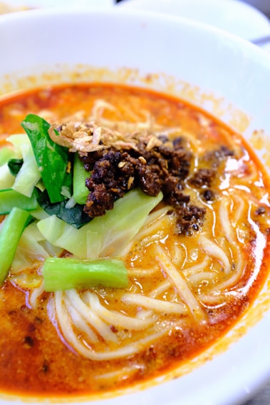 A bowl of spicy noodle soup topped with minced meat, fresh green vegetables, and crispy fried onions. The soup base is rich and reddish-orange, indicating a flavorful and possibly spicy broth.