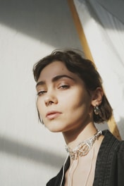 A person with short dark hair and a septum piercing is looking directly at the camera. They are wearing a decorative necklace made of intricate metalwork and a dark textured jacket. Soft light from the side creates shadows on the person's face, enhancing the mood.