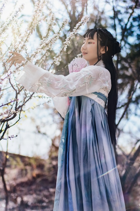 A woman wearing a traditional hanfu dress in shades of blue and white stands near blossoming branches, gently touching the flowers. Her long hair is styled elegantly, and she holds a fan in one hand. The sunlight filters through the trees, creating a soft glow around her.