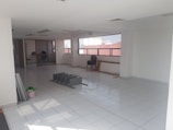 A spacious, empty room features tiled flooring and a few scattered objects. There are chairs near a window and some items like metal shelves and a plastic bag on the floor. The room has a simple and utilitarian design, with large windows allowing natural light to enter.