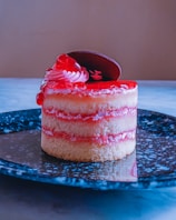 A small, layered dessert featuring sponge cake and red filling, topped with a swirl of cream and a chocolate piece. It is presented on a black speckled plate, set against a neutral background.