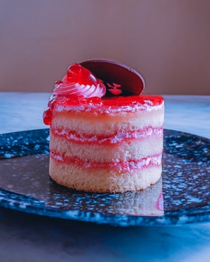 A small, layered dessert featuring sponge cake and red filling, topped with a swirl of cream and a chocolate piece. It is presented on a black speckled plate, set against a neutral background.