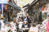 A bustling market street, densely packed with various goods on display. People wearing helmets walk and ride scooters through the narrow path between stalls overflowing with household items, plastic products, and other merchandise.