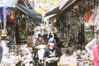 A bustling market street, densely packed with various goods on display. People wearing helmets walk and ride scooters through the narrow path between stalls overflowing with household items, plastic products, and other merchandise.
