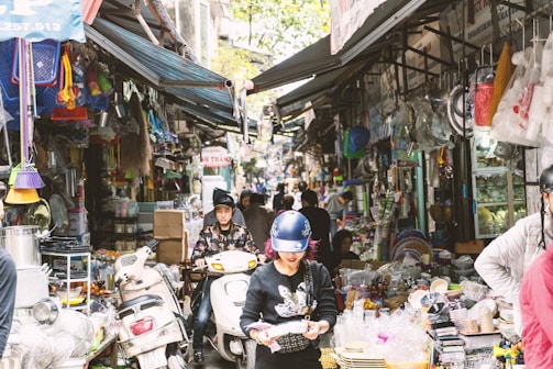 A bustling market street, densely packed with various goods on display. People wearing helmets walk and ride scooters through the narrow path between stalls overflowing with household items, plastic products, and other merchandise.
