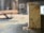 A piece of wood with visible tree rings is positioned upright on a table saw. Sawdust is scattered around, indicating recent cutting activity. The background shows a blurred interior of a workshop or carpentry setting.