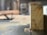 A piece of wood with visible tree rings is positioned upright on a table saw. Sawdust is scattered around, indicating recent cutting activity. The background shows a blurred interior of a workshop or carpentry setting.