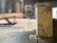 A piece of wood with visible tree rings is positioned upright on a table saw. Sawdust is scattered around, indicating recent cutting activity. The background shows a blurred interior of a workshop or carpentry setting.