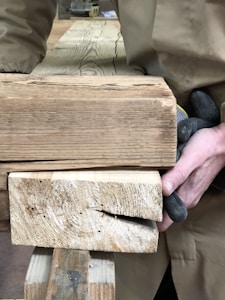 A person wearing gloves is handling several large wooden planks, which appear to be made of rough-sawn timber. The wood has visible grain patterns and texture, indicating a rustic and natural appearance. The individual is wearing a brown coat, and part of their arm is visible.