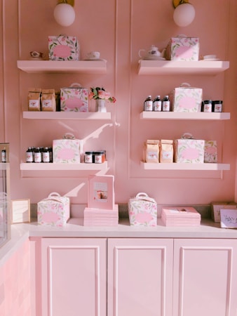 A pastel pink display wall features multiple shelves stocked with elegantly packaged products. Items include floral-patterned boxes, glass jars with dark lids, coffee bags, books, and teacups. A small vase with pink flowers adds a decorative touch, and the overall setting is illuminated with warm light, enhancing the soft color palette.