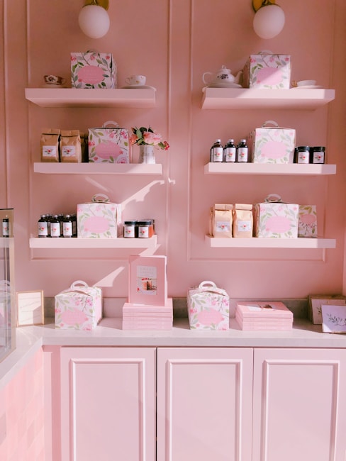 A pastel pink display wall features multiple shelves stocked with elegantly packaged products. Items include floral-patterned boxes, glass jars with dark lids, coffee bags, books, and teacups. A small vase with pink flowers adds a decorative touch, and the overall setting is illuminated with warm light, enhancing the soft color palette.