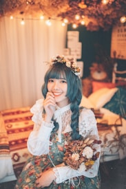 A woman with blue hair styled in braids, adorned with a floral crown. She wears a vintage-style dress with lace details and holds a bouquet of dried flowers. The setting is cozy, with warm string lights and a bed with patterned cushions and a knitted blanket in the background.