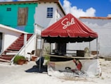 A red tent with the Stoli logo is set up outside, alongside a building with peeling turquoise and white paint. Red wooden stairs lead to a boarded-up door. The area appears somewhat neglected, with a few potted plants, a metal chair, and a wheelbarrow nearby. There's an advertising banner on the tent featuring a person in a red outfit sitting on a zebra illustration.