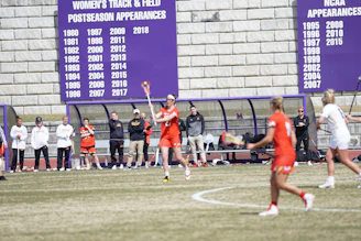 A women's lacrosse game is in progress on a field with players wearing red and white uniforms. The background features a large purple sign listing track and field postseason appearances. Spectators and team members are visible on the sidelines, some standing near a bench area.