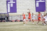 A group of female lacrosse players are actively engaged in a match on a grassy field. Players are wearing athletic gear with some teams in red jerseys and others in white. One player is leaping into the air reaching to catch the ball with a lacrosse stick. In the background, a coach stands near a bench and large purple banners detailing sports achievements are mounted on a stone wall.