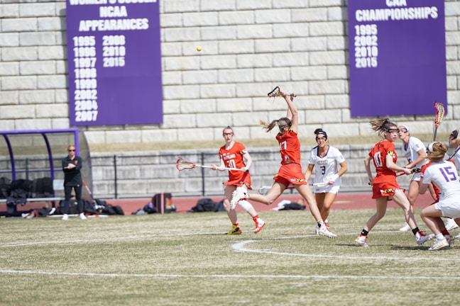 A group of female lacrosse players are actively engaged in a match on a grassy field. Players are wearing athletic gear with some teams in red jerseys and others in white. One player is leaping into the air reaching to catch the ball with a lacrosse stick. In the background, a coach stands near a bench and large purple banners detailing sports achievements are mounted on a stone wall.