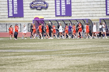 A group of female athletes, wearing red jerseys, walk in a line on a sports field. They appear to be part of a lacrosse team. The background features a stone wall with banners displaying national championship years. Nearby, there is a purple and yellow covered bench area.