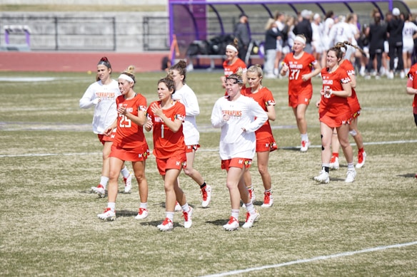 A group of female athletes wearing red and white uniforms are jogging on a grassy sports field. The players are part of a team, with some in red jerseys and others in white tops. They seem to be warming up or cooling down, and there is a bench with people in the background.