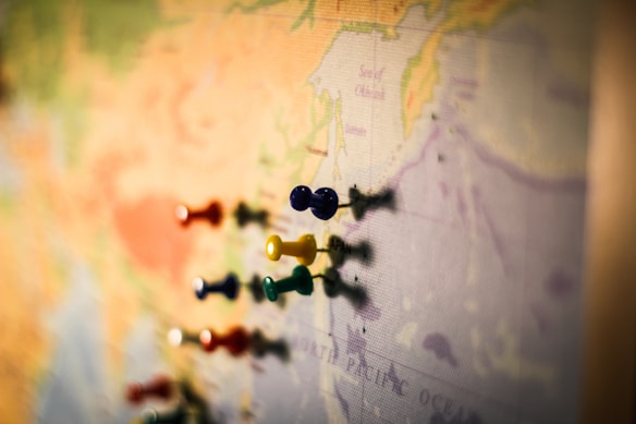Various colored push pins placed on different locations of a world map, indicating marked points of interest or visited places. The focus on the push pins creates a sense of three-dimensional depth against the backdrop of the map.