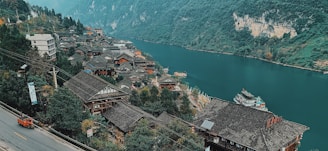A village with traditional wooden houses is nestled along the banks of a serene river. The scene is framed by lush green mountains and a cloudy sky. A road runs along the side of the village with a small orange vehicle parked at the edge. There are several boats docked near the riverbank, and some buildings have modern elements such as solar panels.