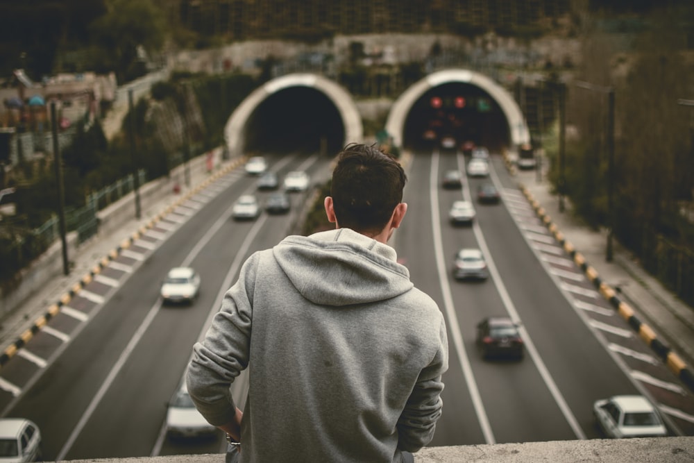 man wearing gray hoodie standing above road with cars running during daytime
