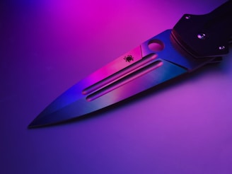 A close-up view of a knife with a metallic blade featuring a subtle spider logo. The blade reflects vibrant neon colors, creating a gradient effect of purples and blues. The background is softly illuminated, enhancing the colors on the knife.